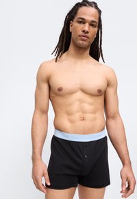 Νεαρός άνδρας με dreadlocks που φοράει μαύρα εσώρουχα Calvin Klein με ανοιχτό μπλε λάστιχο στη μέση, στέκεται μπροστά σε απλή φόντο.