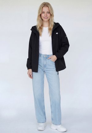 Fekete, könnyű dzseki állítható kapucnival, fehér emblémával a mellkason, fehér póló és világoskék, magasderekú farmernadrág felett viselve, fehér tornacipővel párosítva.