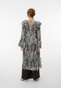 Femme aux cheveux relevés en chignon, portant une robe longue transparente à motifs avec des volants, par-dessus un pantalon large noir, et des chaussures crème à talons bas, vue de dos.