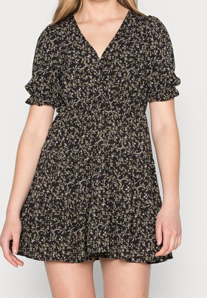 Schwarzes kurzes Kleid mit kleinem lila Blumenmuster, V-Ausschnitt, aufgeblähten kurzen Ärmeln und gestuftem Rock, getragen von einer stehenden Frau.