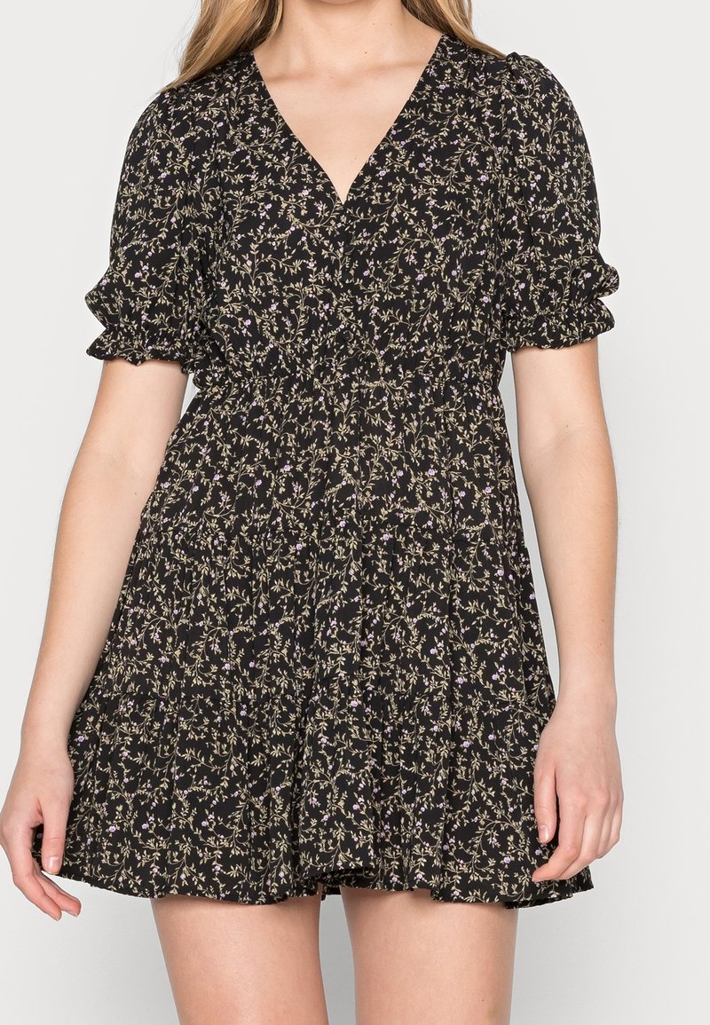 Vestito corto nero con piccola stampa floreale viola, scollo a V, maniche corte a sbuffo e gonna a balze indossato da una donna in piedi.