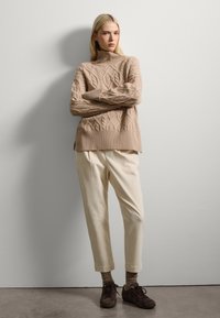 Jersey de punto beige con cuello alto, combinado con pantalones color crema y zapatos marrón oscuro. Textura suave y corte relajado.