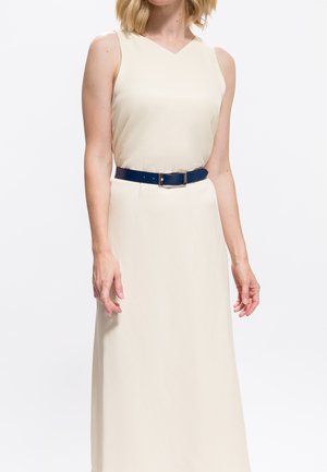 Donna che indossa un vestito senza maniche color crema con scollatura a V e una cintura blu scuro in vita, su uno sfondo bianco.