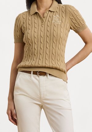 Femme portant un polo beige à manches courtes en maille torsadée avec initiales brodées, pantalon blanc et ceinture marron sur un fond uni.