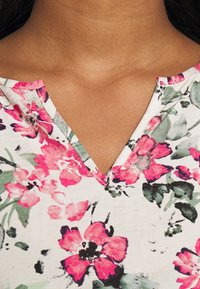 Bluzka w kwiaty z dekoltem w serek, z białym tłem i różowo-zielonymi kwiatami, wykonana z lekkiego materiału o gładkiej fakturze.