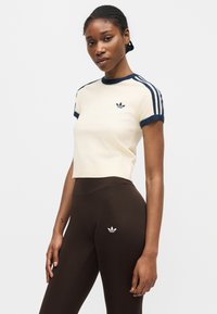 Γυναίκα που φοράει κρεμ cropped t-shirt με ναυτικό μπλε ρίγες και καφέ leggings, και τα δύο με λογότυπα Adidas trefoil, σε απλό λευκό φόντο.
