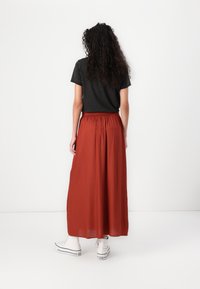 Jupe longue couleur rouille avec une taille élastique, assortie à un t-shirt noir court et des baskets blanches. Le tissu semble lisse et léger.