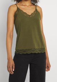 Femme portant un haut sans manches olive avec des bordures en dentelle et des bretelles spaghetti, accompagnée de pantalons noirs, se tenant devant un fond uni.