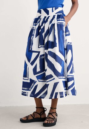 Una falda blanca hasta la rodilla con un audaz patrón geométrico azul, con una cintura elástica y bolsillos laterales. Llevada con sandalias negras de tiras.