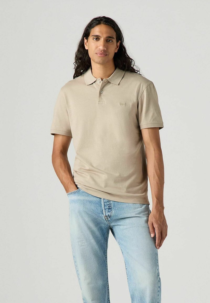 Beige pikétröja med krage, korta ärmar och en diskret logotyp. Bärs med ljusblå jeans som har en avslappnad passform och en urtvättad look.