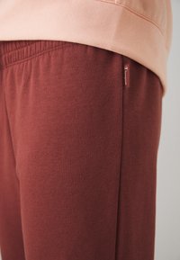 Pantalones de chándal color burdeos en una tela suave, con cintura elástica y etiqueta de logo visible en el costado. Textura suave con un ajuste relajado.