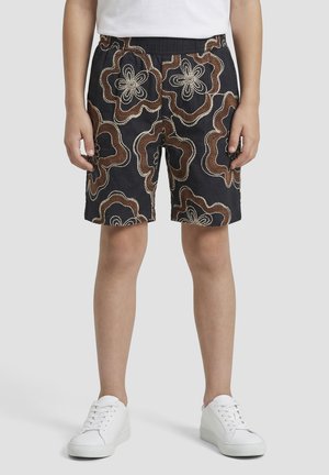 Crne kratke hlače do koljena s velikim smeđim i bež vezanim cvjetnim uzorcima koje nosi stojeća osoba u bijelim tenisicama.