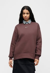 Μπορντό φούτερ με λαιμόκοψη, ριμπ μανσέτες και λογότυπο Nike σε ασημί χρώμα. Φορεμένο πάνω από ένα ανοιχτό μπλε πουκάμισο με γιακά. Ομαλή υφή υφάσματος.