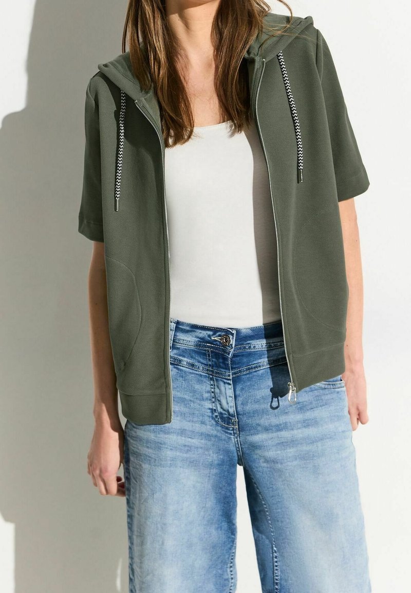 Femme portant un sweat à capuche zippé manche courte vert olive par-dessus un débardeur blanc et un jean taille haute bleu clair sur un fond uni.
