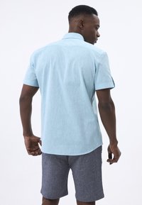 Hellblauses Kurzarmhemd aus strukturiertem Stoff, mit klassischem Kragen und hochgerollten Ärmeln, kombiniert mit dunkelgrauen Shorts.