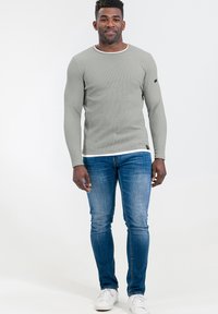 Graues langärmliges Ripp-T-Shirt mit weißem Ausschnitt, getragen mit blauen Slim-Fit-Jeans und weißen Sneakers. Das Shirt hat ein kleines Logo am Ärmel.