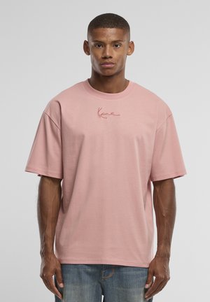 Un homme se tient face à l’avant, portant un T-shirt rose ample avec un petit logo brodé sur la poitrine et un jean bleu sur un fond gris uni.