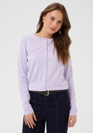 Kvinde med langt brunt hår iført en lyse-lilla knappet cardigan, mørke jeans, sølvhalskæde og øreringe mod en ensfarvet lys baggrund.