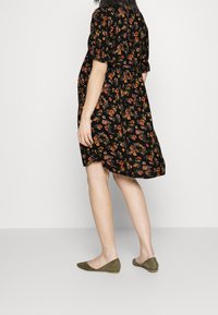 Schwarzes Blumenkleid mit kurzen Ärmeln und einer A-Linie-Silhouette, das orangefarbene, gelbe und grüne Muster aufweist. Kombiniert mit olivgrünen Ballerinas.