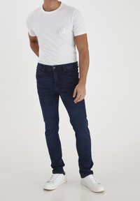 Vit kortärmad t-shirt med en reflekterande fyrkantig design, tillsammans med mörkblå slim-fit jeans och vita sneakers.