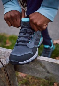 Szare buty sportowe z czarnymi i turkusowymi akcentami; charakteryzują się teksturowaną siateczką, zapięciem na sznurówki oraz wspierającą podeszwą.