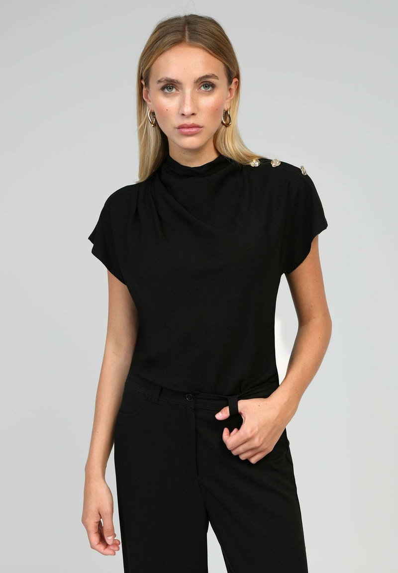 Haut noir à manches courtes avec un col montant, un design drapé et trois boutons décoratifs à l'épaule. Tissu lisse avec un subtil éclat.