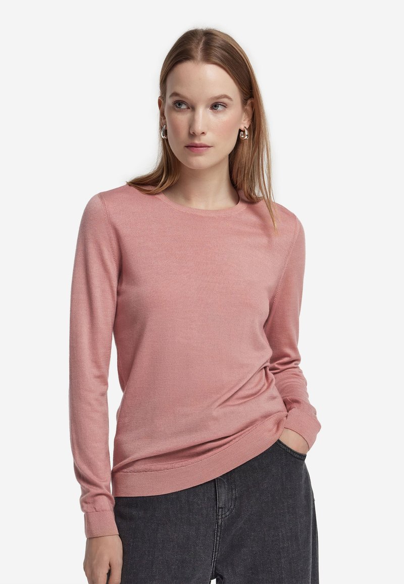 Kvinde med langt, glat hår har en pink langærmet sweater og mørke jeans på, kigger let til siden med den ene hånd i lommen.