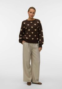 Hnědý svetr s béžovými vzory srdcí, spárovaný se světle béžovými širokými kalhotami. Jednoduchý design s žebrovanými manžetami a lemem. Hnědé boty.