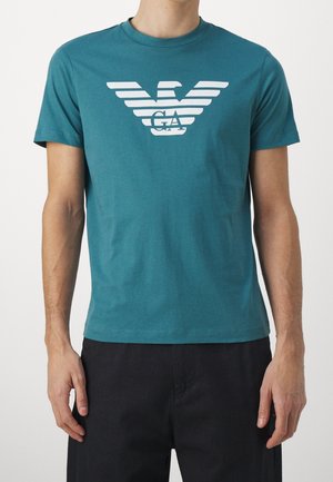 Homme portant un t-shirt à manches courtes bleu sarcelle avec un logo d'aigle stylisé blanc et les lettres GA sur la poitrine, associé à un pantalon noir, sur fond uni.