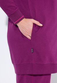 Haut de sport violet avec un col et des poignets côtelés. Une main est placée dans une poche latérale. Présente une petite étiquette de logo sur l'ourlet.