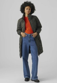 Lange olijfgroene donsjas met een hoge kraag, over een felrood shirt, gecombineerd met hoge taille blauwe jeans en zwarte schoenen.