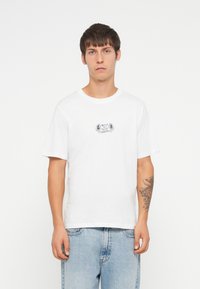 Hvit t-skjorte i bomull med korte ermer, rund hals og et lite svart grafisk design på brystet. Brukt med lyseblå denim jeans.