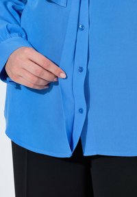 Camicia azzurro chiaro a maniche lunghe realizzata in tessuto morbido, con chiusura a bottoni frontale, orlo arrotondato e una sottile trama. Una mano tiene il tessuto.
