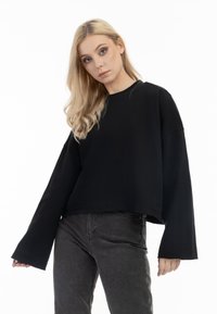Sweatshirt negru scurt cu mâneci lungi și largi. Textură moale și design simplu. Purtați-l cu blugi gri închis. Poziționat pe un fundal alb.