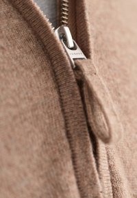 Brauner Strickpullover mit einem silbernen Reißverschluss und Markenetikett, der eine weiche Textur zeigt und eine Nahaufnahme des Reißverschlussdetails präsentiert.
