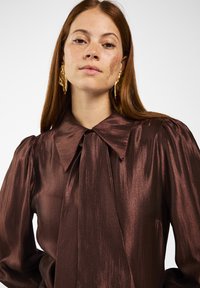 Bruine satijnen blouse met een grote kraag, geplooide schouders en een strik rond de hals. Beschikt over een gladde, glanzende textuur en aansluitende mouwen.