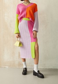 Dzianinowy sweter w kolorach pomarańczowym, różowym i zielonym z dekoltem w serek, zestawiony z pasującą spódnicą z bocznym rozcięciem. Dopełniony żółtą torebką.