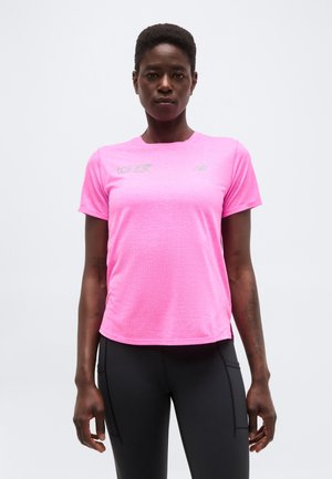 Γυναίκα που στέκεται με το πρόσωπο προς τα εμπρός, φορώντας φωτεινό ροζ αθλητικό μπλουζάκι με κοντά μανίκια και μαύρο κολάν, με φόντο ένα απλό φωτεινό περιβάλλον.