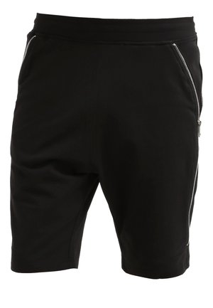Schwarzes V-Ausschnitt-T-Shirt, schwarze Sportshorts mit Reißverschlussdetail und schwarze Sneakers. Trägt eine schwarze Reisetasche mit grauen Griffen.