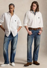 Zwei Models tragen weiße Hemden mit NY-Logos und blaue Jeans; eines trägt weiße Turnschuhe, das andere braune Stiefel mit Gürtel und Schal.