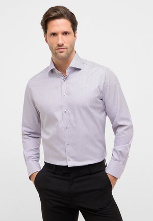 Uomo che indossa una camicia a maniche lunghe di colore chiaro, con motivi, pantaloni eleganti neri, mani in tasca, in piedi davanti a uno sfondo semplice.
