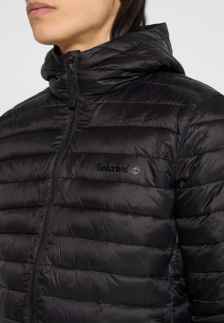 Osoba nosząca czarną pikowaną kurtkę Timberland z kapturem i przednim zamkiem, pokazana od ramion do podbródka.