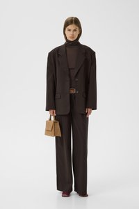 Sieviete, tērpusies pārmērīgi lielā tumši brūnā kostīmā ar platām biksēm, turtleneck, sarkanais apavu pāris un tur rokās mazu gaiši brūnu rokassomu.