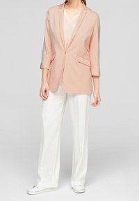 Vaaleanpunainen bleiseri viistokauluksella, kahdella etutaskulla ja puolipitkillä hihoilla, yhdistetty valkoisiin leveälahkeisiin housuihin ja valkoisiin liukusandaaleihin.