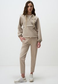 Felpa beige con cappuccio e dettagli dorati, abbinata a pantaloni slim-fit dello stesso colore e sneaker bianchi con dettagli beige. Tessuto morbido e vestibilità comoda.