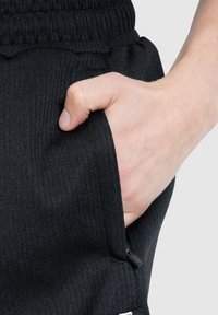 Svarta ribbade joggers med elastisk midja och en sidoficka. En hand vilar i fickan och framhäver tygtexturen.