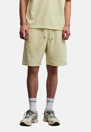Person trägt blassgelbe Shorts und passendes T-Shirt, weiße Socken mit dem Text "PEGADOR" und cremefarbene Sportschuhe, steht aufrecht.
