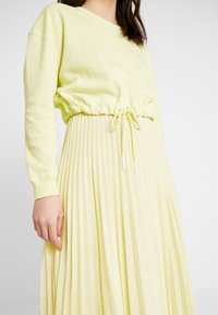 Felpa gialla chiara con vita arricciata e cordoncino, abbinata a una gonna leggera e plissettata gialla chiara, che mette in mostra una morbida texture e un design informale.