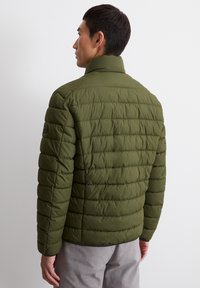 Olive green puffer jacket with horizontal stitching, stand collar, and a smooth texture. Features a logo detail on the left sleeve.