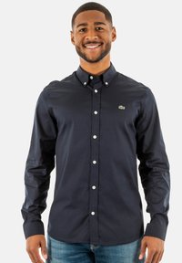 Camicia button-up di un blu navy con colletto classico, bottoni bianchi e un piccolo logo di coccodrillo verde sul petto. Tessuto a trama liscia.